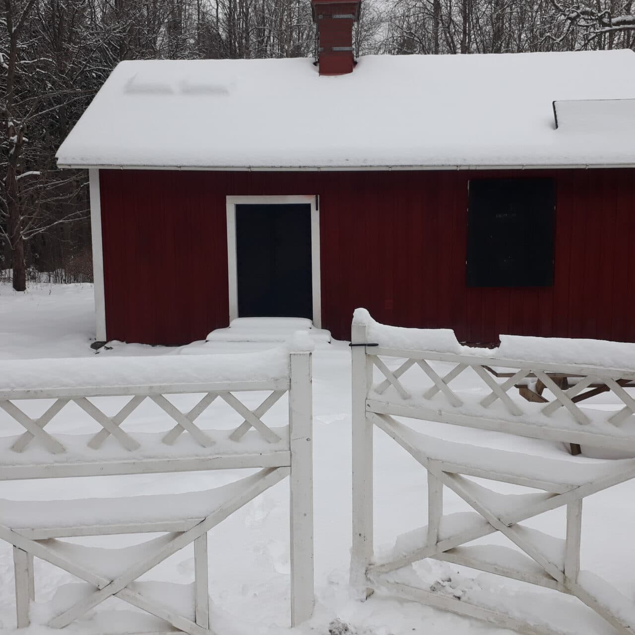 Vinterbild av Lillåstugan med snö på taket och den snöklädda grinden i förgrunden.
